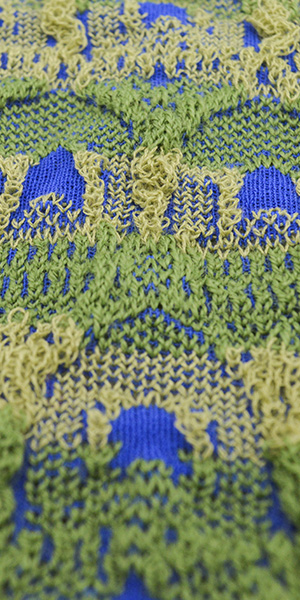 Knit fabric where the textured surface in alternating stripes of darker and lighter greens is accented by spots of deep blue, of which smaller points weave inbetween the greens.