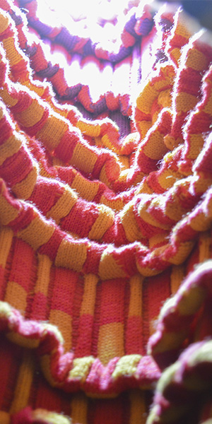 Detail of a neon colored fabric in an alternating checkerboard pattern in pink, purple and yellow. The surface wavers with numerious pleats illuminated by light from above.