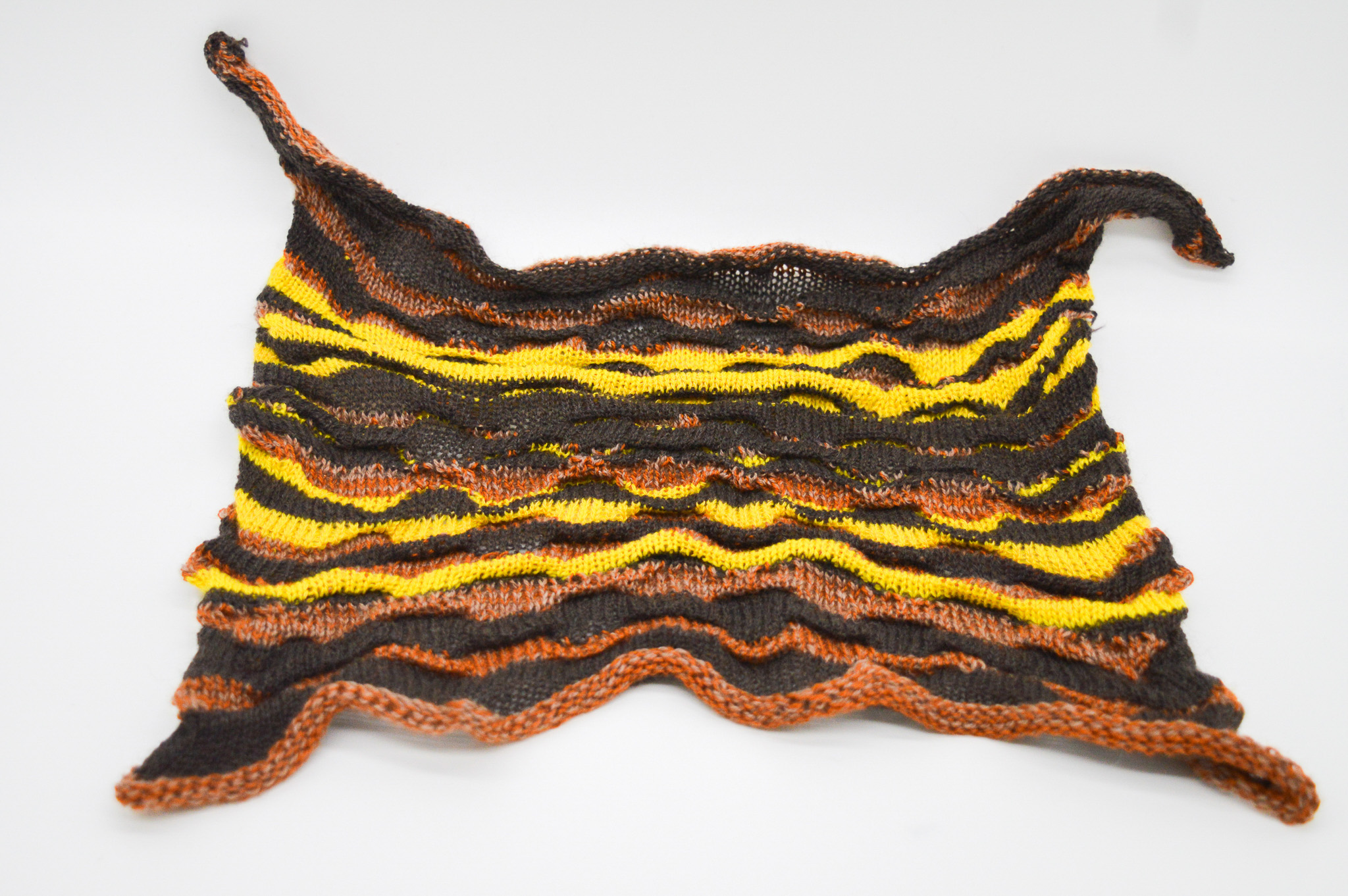 Loosly rectangular knit sample with pulled out corners whose textured surface features raised waving stripes of yellow, black and brown.