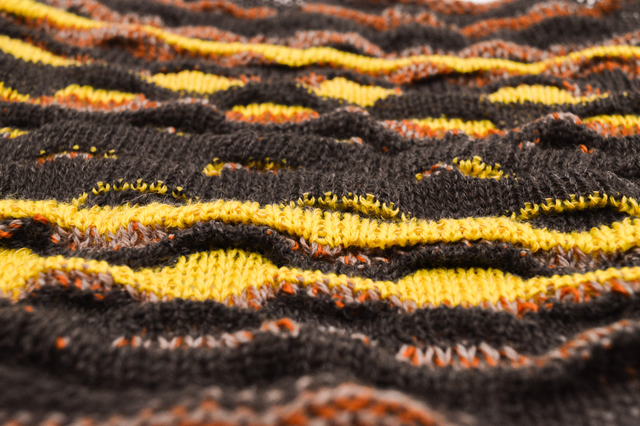 Closeup of a knit fabric with a textured surface made up of raised waves in varying alternating stripes of black, brown and yellow.