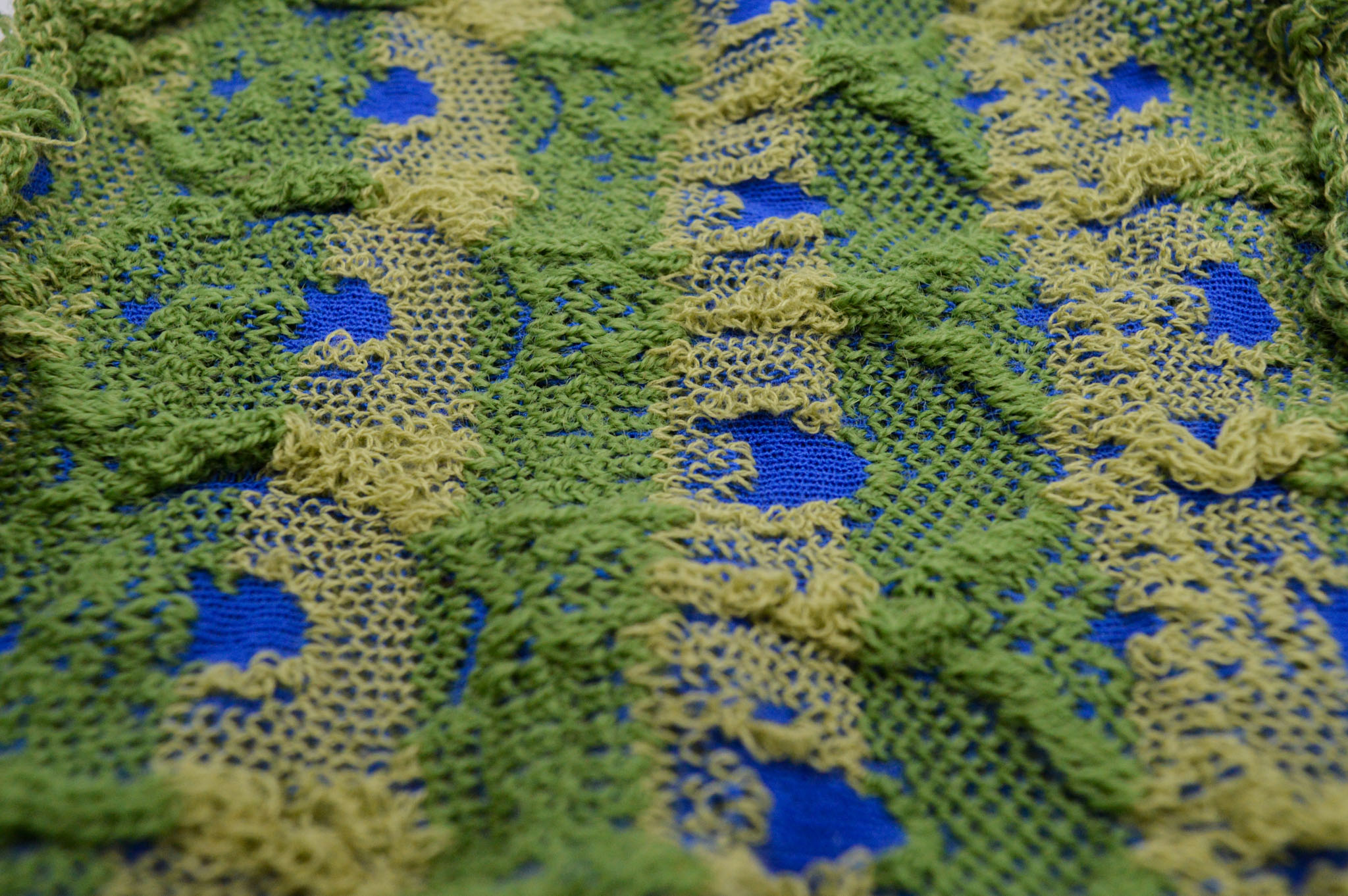 Closeup of a horizontal fabric where the surface is textured in an arch pattern, in alternating stripes of darker and lighter greens accented by spots of deep blue of which smaller points weave inbetween the greens.