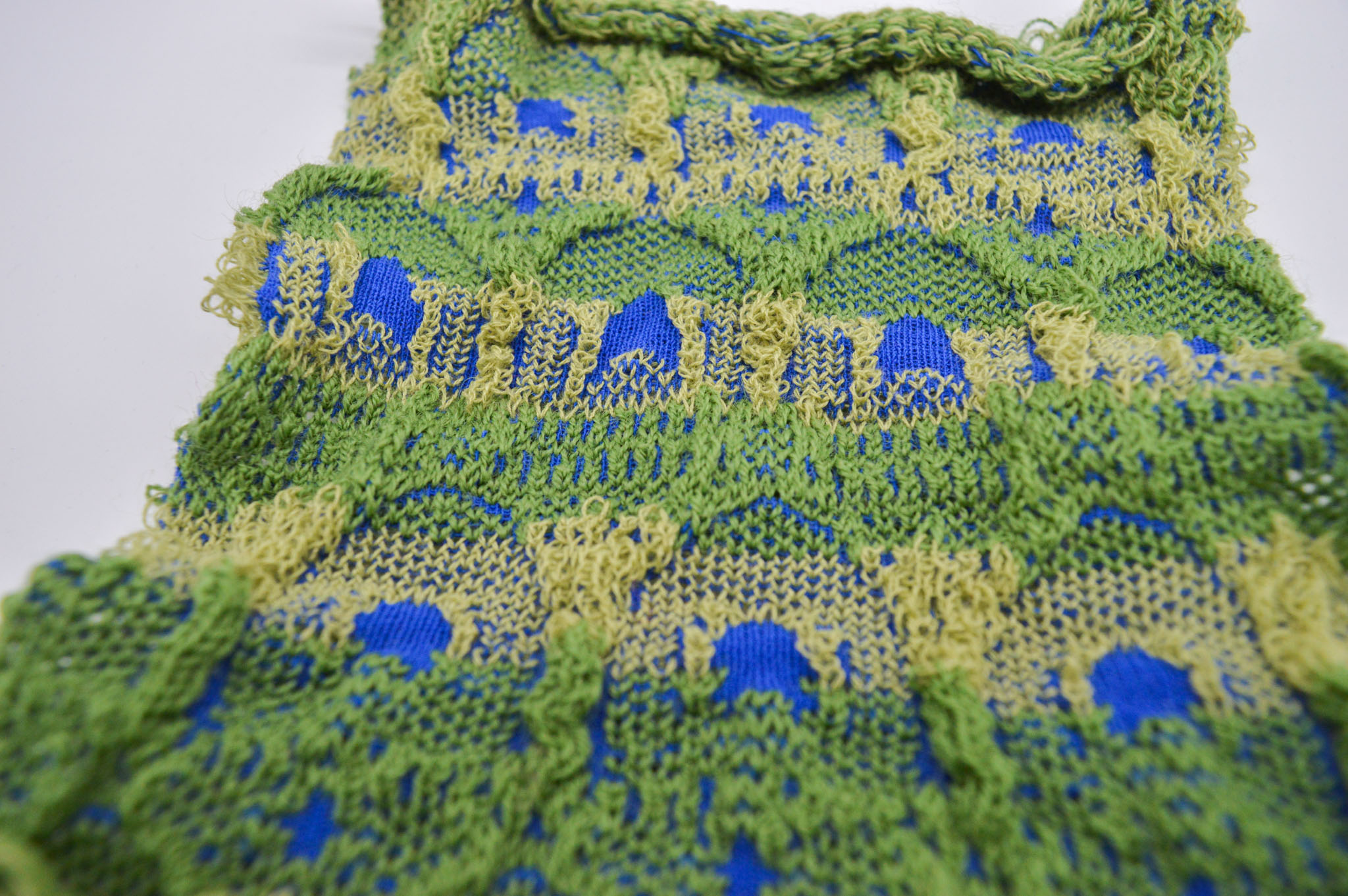 Vertical knit green fabric with blue accents on a white background which features an alternating arch pattern across the surface made of raised texture which frames areas of interlocked blue and green stitches around a central blue point. The top edge curls inwards.