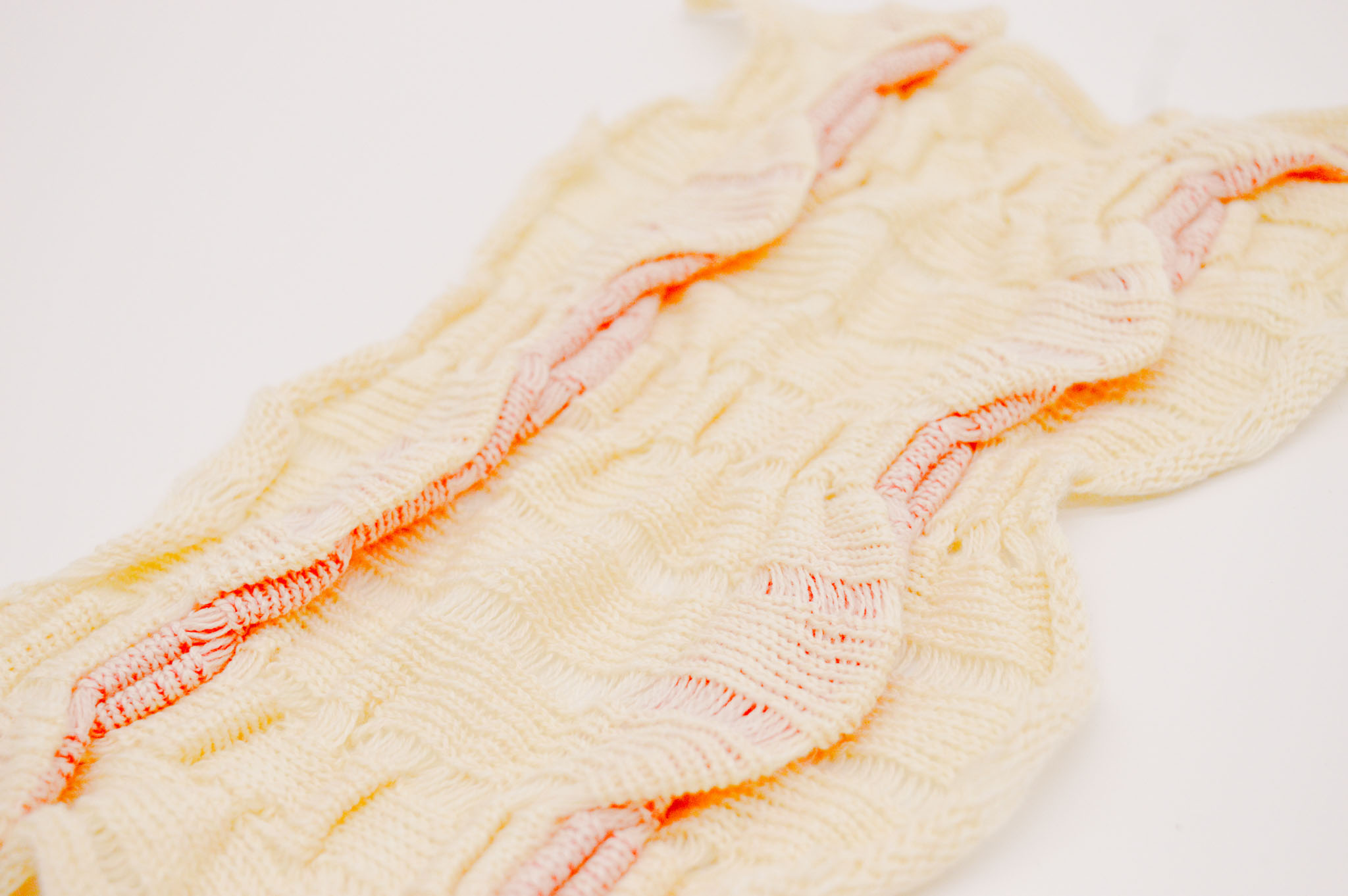 Cream colored rectangular knit fabric on a light background at an angle. The surface is made up of a series of pleats and folds in a textured wave pattern, with orange accents peeking out from underneath the largest folds, casting saturated orange shadows.