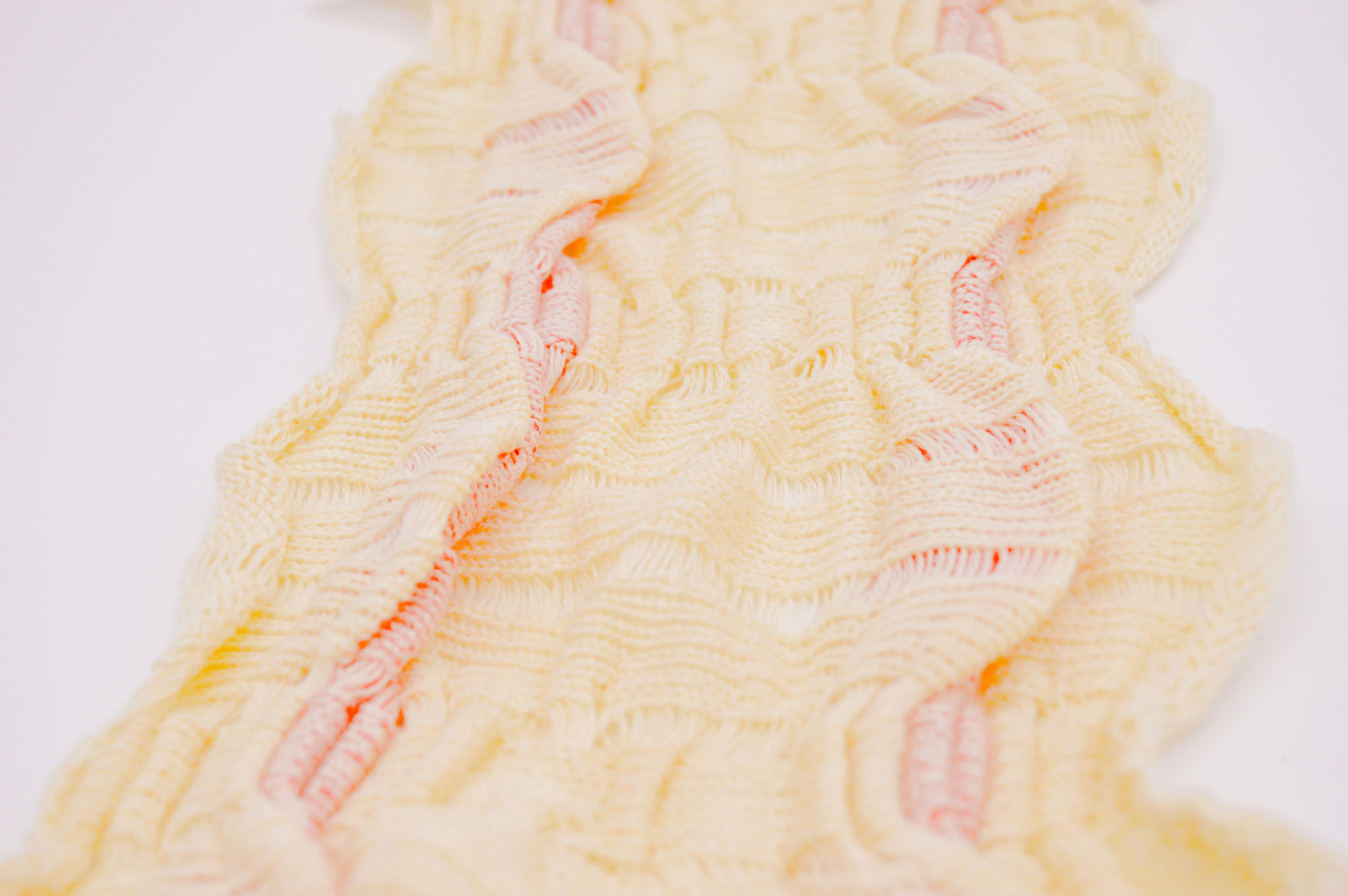 Cream colored knit fabric on a light background made up of a series of pleats and folds in a wave pattern to create a textured surface, with orange accents peeking out from behind the spaces between the knitting.