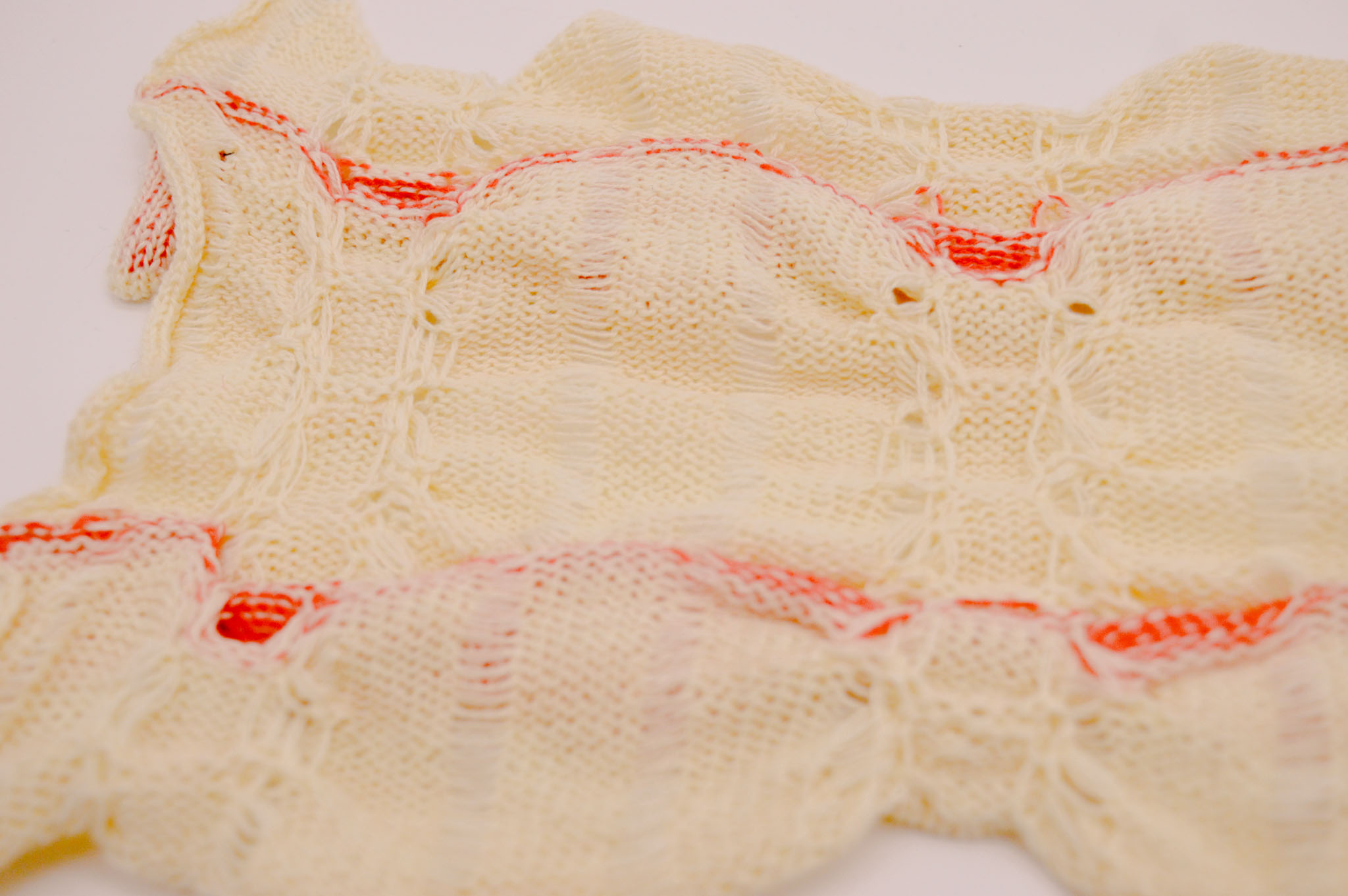 Detail of the wrong side of a cream colored knit fabric on a white background with a wavering surface, which shows two columns of pulled in stitches crossed by two bright orange stripes.