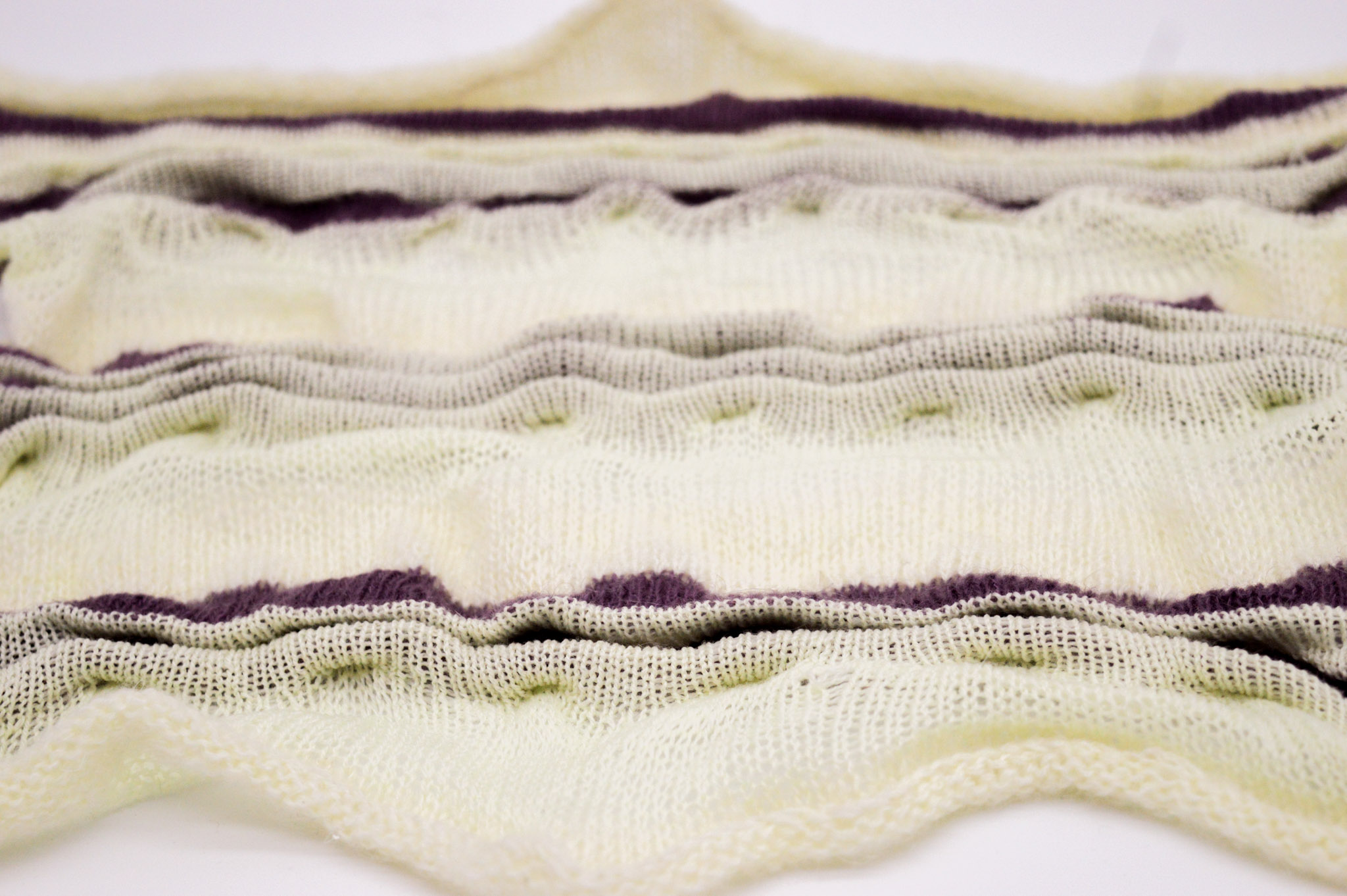 Close view of a horizontal white knit fabric with pleated waves striped in purple and white with ripples where the pleats rise out of the fabric and the center of the edges extend out into slopes.