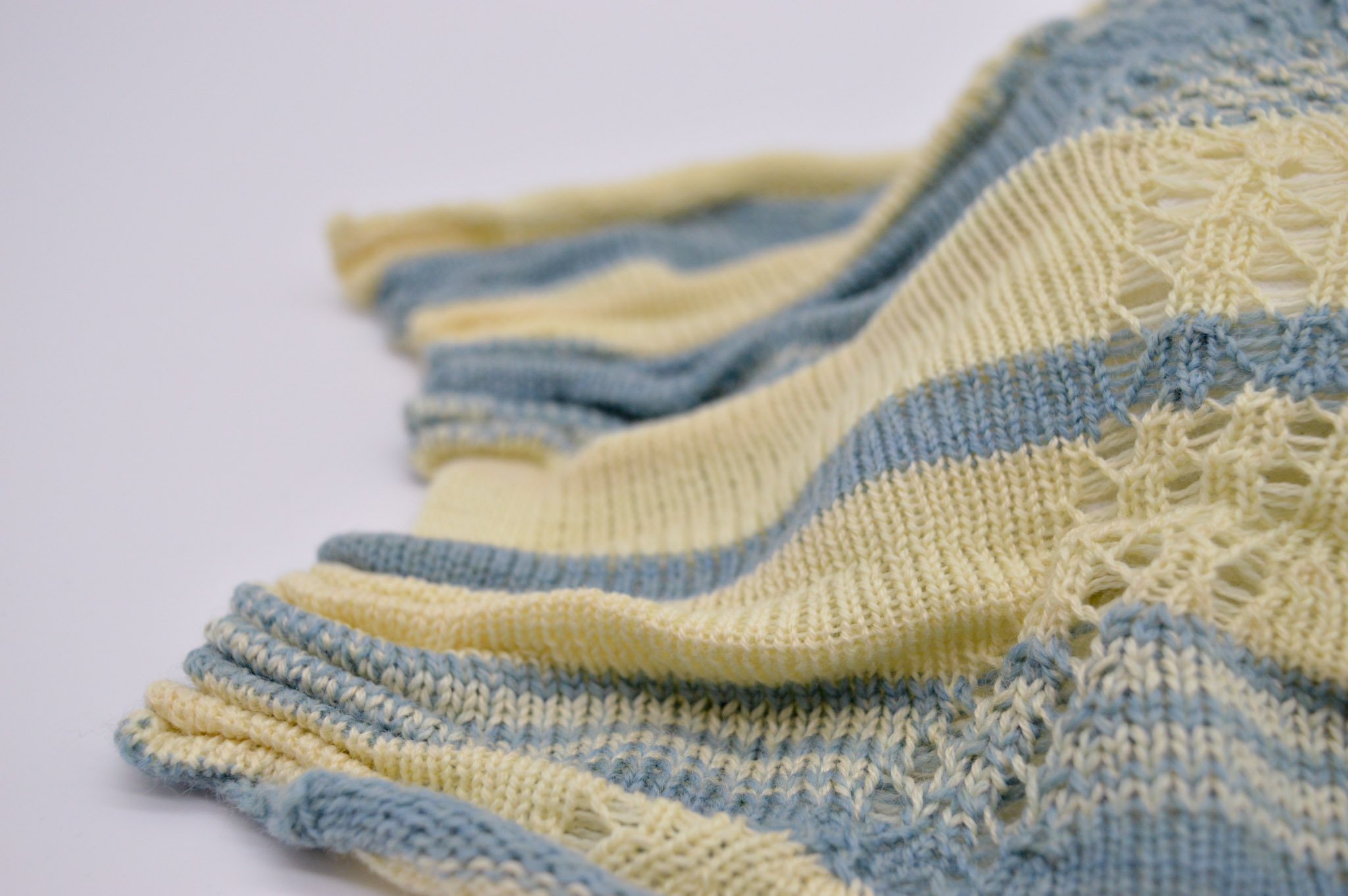 Close-up of a knit fabric on a white backdrop with blue and cream stripes and lacey holes accenting the fabrics shape around a spherical form. The left edge ofthe fabric going into the background shows groupings of tight pleasts that causes the folds and form.