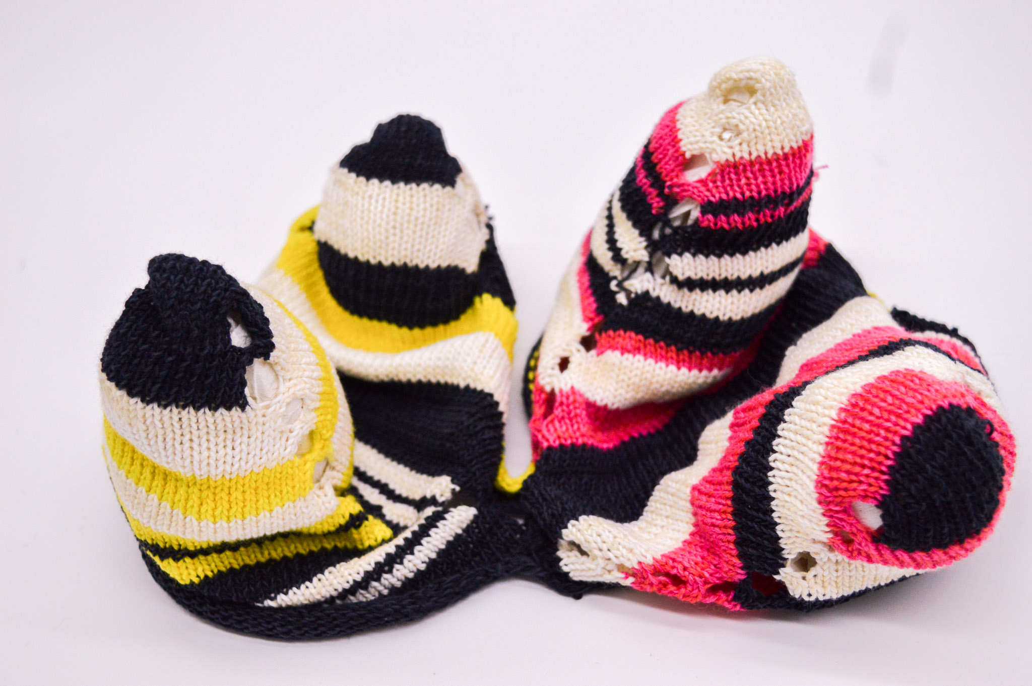 Knit sample with a soft structure in the form of four symmetrical cones on a white backdrop. The cones have contrasting striping in black and white, with the cones to the left featuring neon yellow stripes while the ones to the right feature pink stripes. There are lace holes running up the sides of the sample.