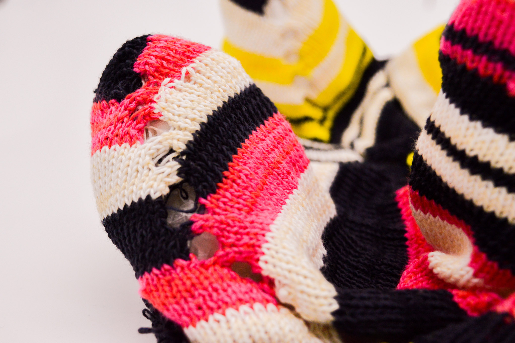 Close-up of a series of soft knit cones whose surface features large contrasting stripes in black and white, with the forground cones additionally having pink striping and the background cones having yellow stripes. Openwork holes run down prominent side of the cone in the forground.