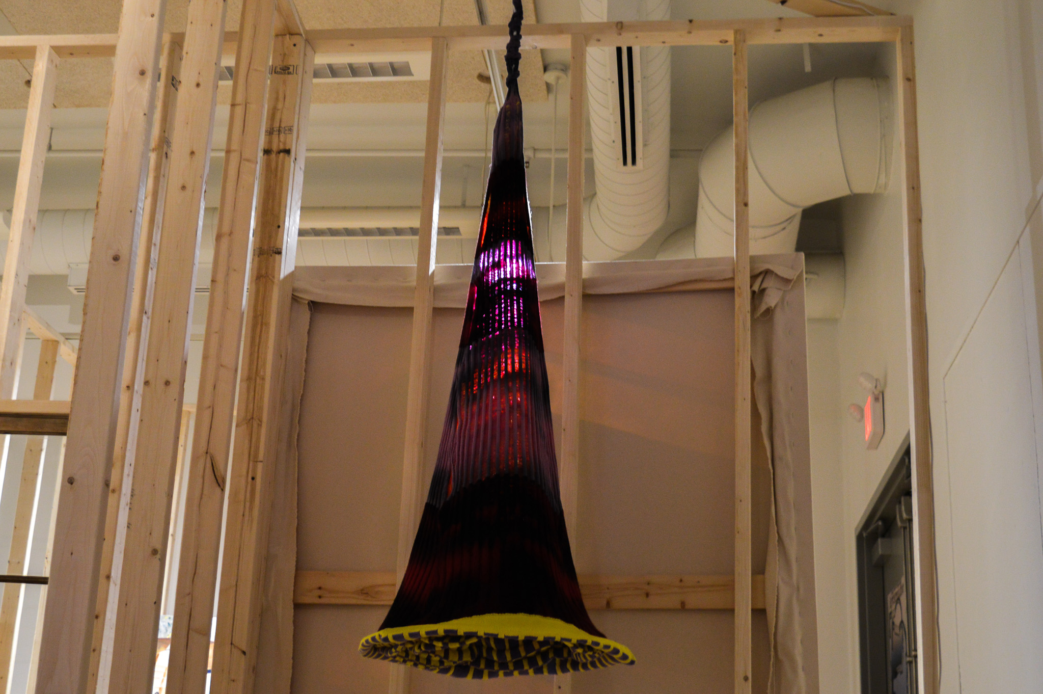 A hanging cone-shaped sculpture surronded by vertical wooden beams and lit from behind. The sculpture is made of knitted fabric in shades of pink, red and black bordered by bright yellow on the bottom, with a checkerboard pattern from inside peeking out. A glow emanates from the inside around the middle, illuminating the knit structure.