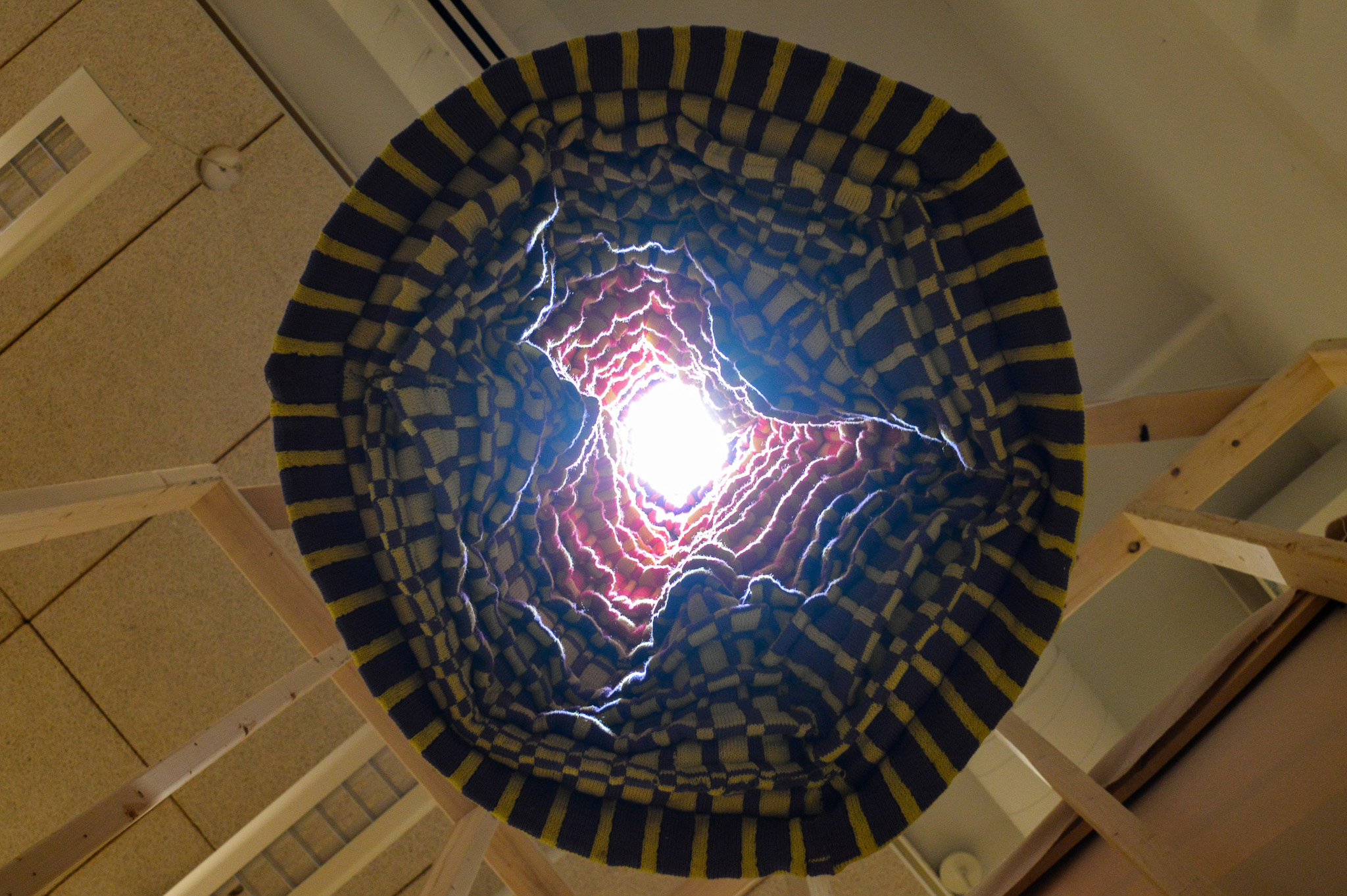 View straight up into a hanging circular sculpture made of knit fabric which has a cold light at the top, illuminating the numerous pleats which make up the textured surface of the interior. The bottom features a varied checker pattern in bright purple and yellow, which then transitions into yellow and pink as it goes up into the light.