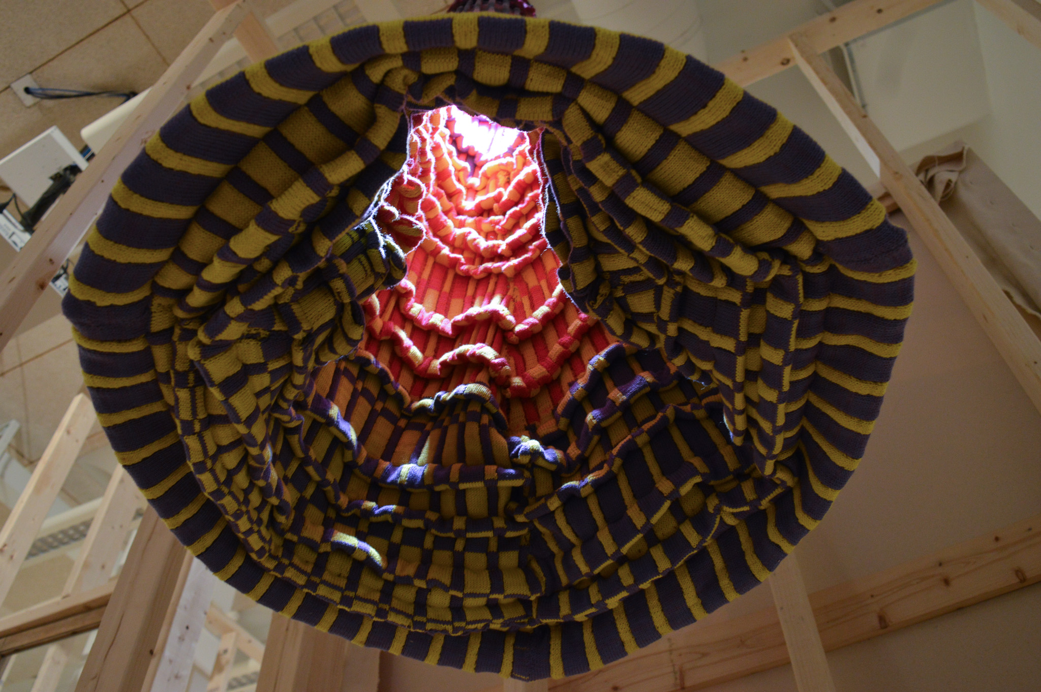 Upward facing view of a hanging circular sculpture at an angle, emphasizing the form created by the many stacked pleats and checkerboard pattern of the bottom. Up further in the sculpture, a bright white light spills out illuminating the neon yellow and pink checkerboard pattern which make up the upper half of the interiors pleats.