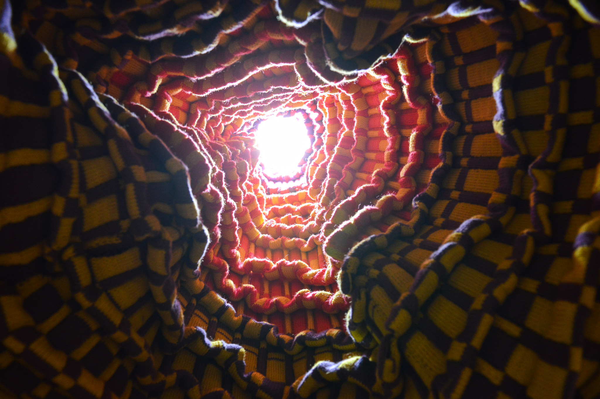 Upward view of a tunnel made of knitted fabric ending in a bright white light that illuminates the forms made by pleats. The walls have a checkerboard pattern in purple and yellow which becomes pink and yellow, and numerous pleats which follow the wavering surface all the way up.