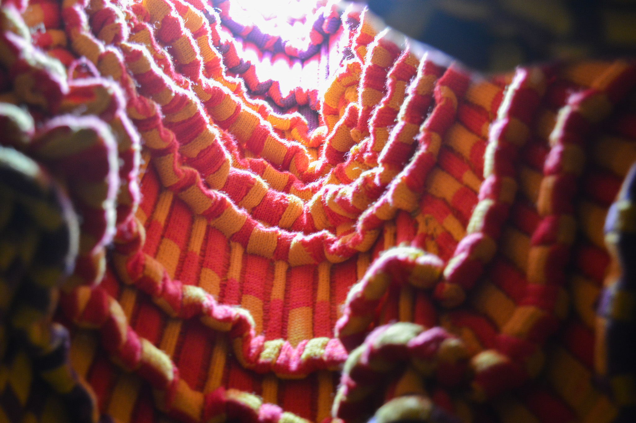 View of a knit fabric with many pleats and a wavering, undulating surface which leads up into a bright white light at the top that illuminates the neon pink and yellow that make up a checkerboard pattern. 