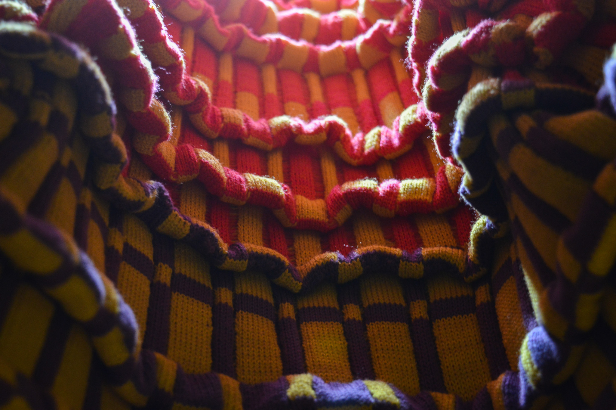 Close view of a brightly colored neon knit fabric with a checkerboard pattern and undulating, wavering surface filled by numerous pleats. The surface is lit with a bright white light from above, the colors starting with purple and yellow and transitioning into pink and yellow.